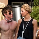 Two young people, one shirtless with sunglasses, the other in a black dress, talking outdoors.