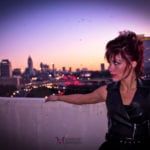 A woman with red hair leaning on a balcony at sunset overlooking a city skyline.
