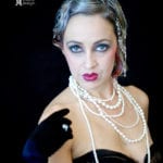 A woman with dramatic makeup and pearl necklaces against a dark background.