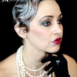 A woman with styled grayish hair, dramatic makeup, and pearl necklace.