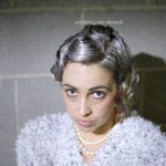A woman with gray braided hair and pearl necklace looks up at the camera.