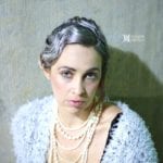 A woman with curled hair wearing a pearl necklace and a light blue shawl.