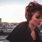 A woman with short red hair and dramatic makeup on a rooftop at sunset.