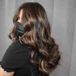A woman with long, wavy brown hair wearing a black mask and black shirt.